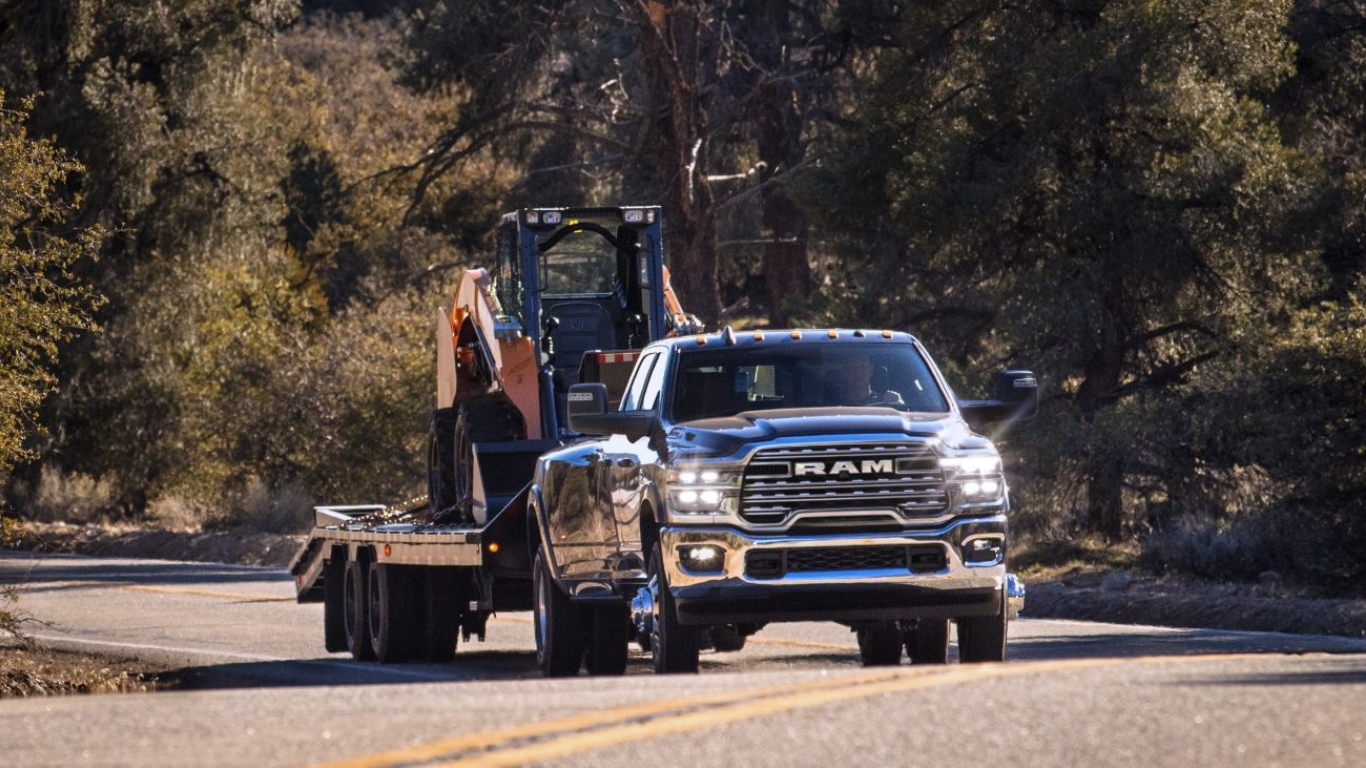 A 2025 Ram 3500 commercial truck pulls a trailer with a bulldozer on it down a county road.