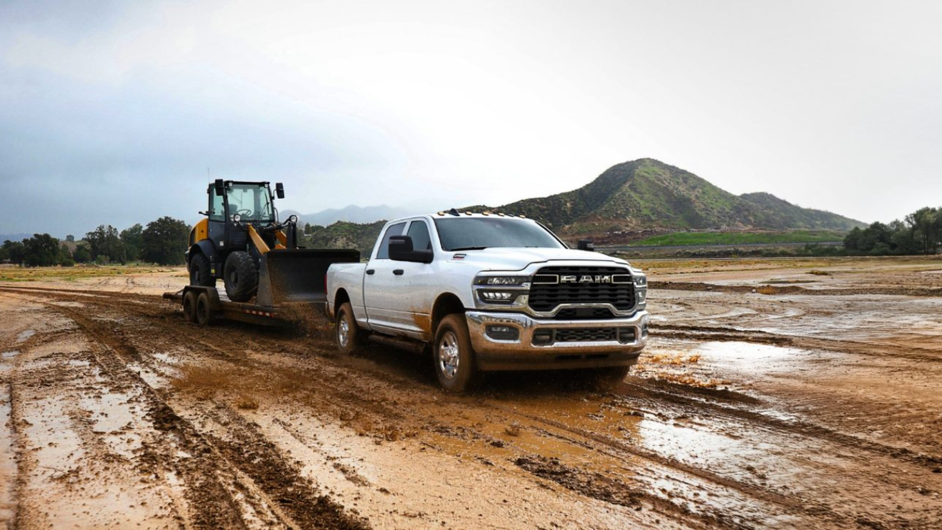 A 2025 Ram 2500 pulls a trailer with a bulldozer on it across a muddy field.